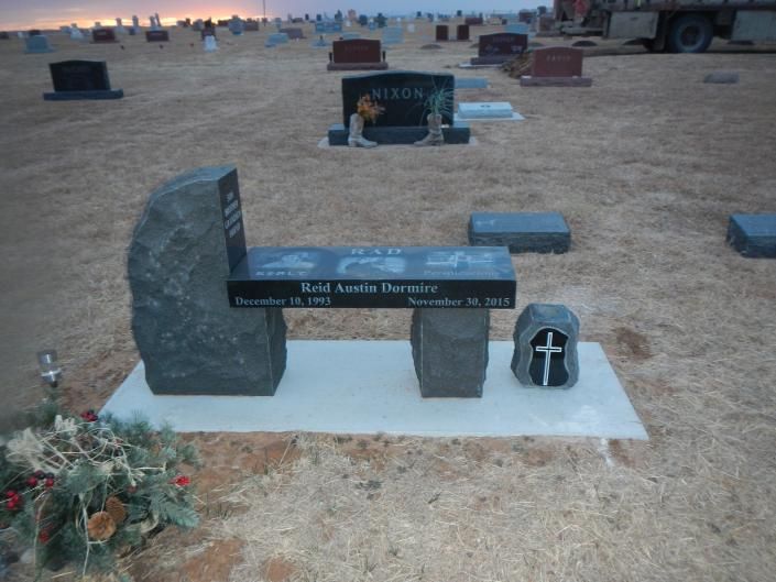Gravestone memorial in a sandy cemetery with flowers and a small cross marker at sunset