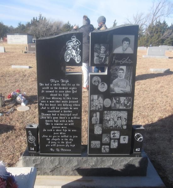 Black memorial headstone with engraved photos and text in a cemetery, with flowers nearby