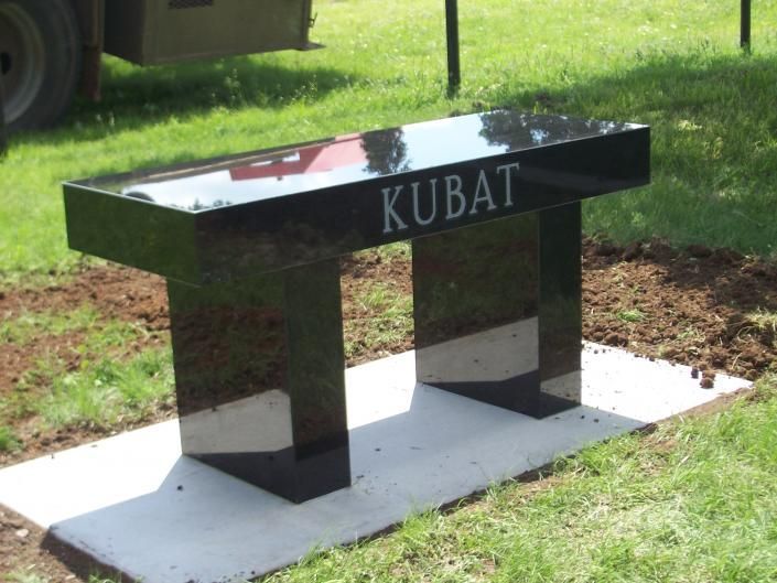 Black granite memorial bench engraved “KUBAT” on a white base in a grassy park area