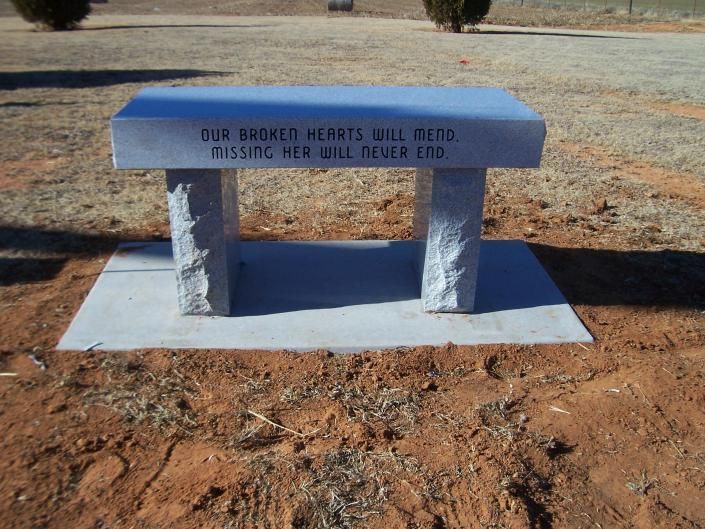 Blue memorial bench on a concrete slab in a dry, grassy outdoor area with engraved text on the backrest