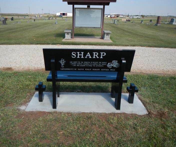 Black memorial bench labeled “SHARP” at a cemetery with graves and a small shelter behind it.