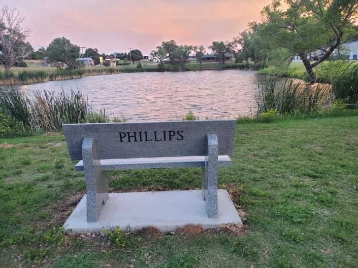 Bench labeled “PHILLIPS” by a grassy lakeside at sunset