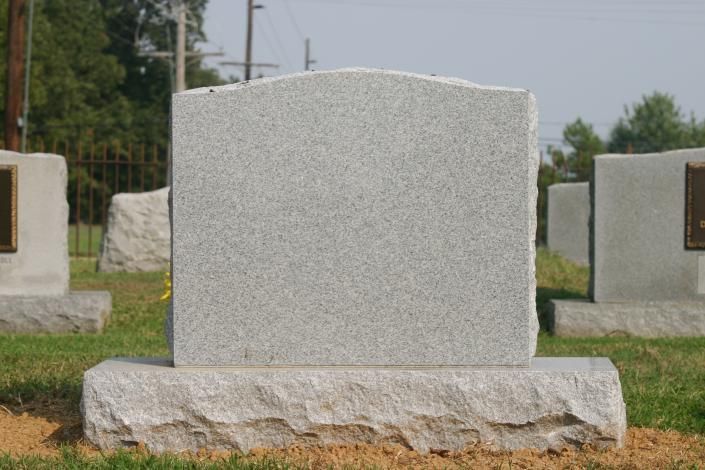 Gray granite gravestone in a cemetery, centered on a stone base with other markers in the background