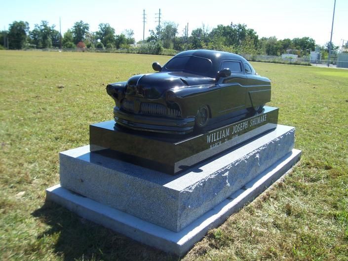 Black sports car monument on a stone pedestal in a grassy field under a clear sky