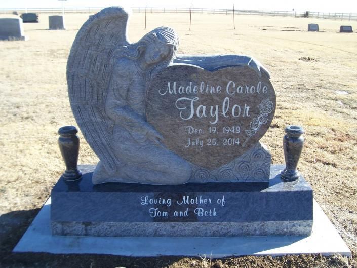 Engraved dark stone memorial headstone in a sandy cemetery, reading “Jaylor” with decorative angel wings.