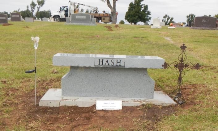 Gray cemetery headstone marked “HASH” on a grassy hillside, with a small cross nearby.