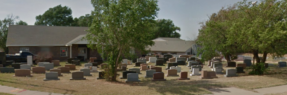 Cemetery with rows of gravestones beneath trees beside a building on a sunny day