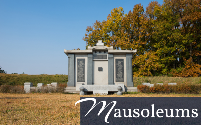 Small gray mausoleum in a grassy cemetery, framed by autumn trees under a clear blue sky.