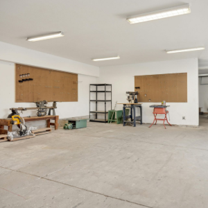 Empty white workshop with concrete floor, boarded windows, and tools along the walls