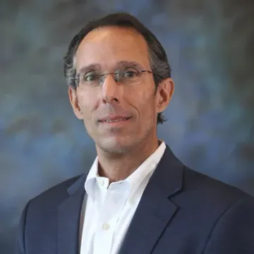 A portrait of a smiling person with shoulder-length, dark hair, wearing a tan shirt against a white background.