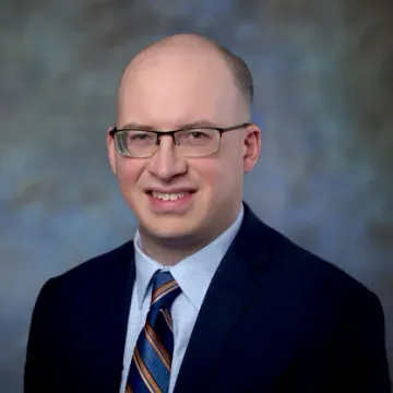 A bald person wearing dark-rimmed glasses smiling, centered against a plain white background.