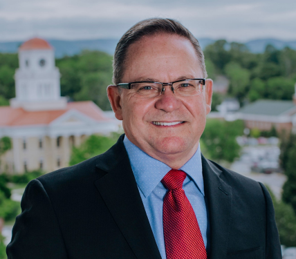 Smiling man in a dark suit and red tie with a blurred campus building in the background