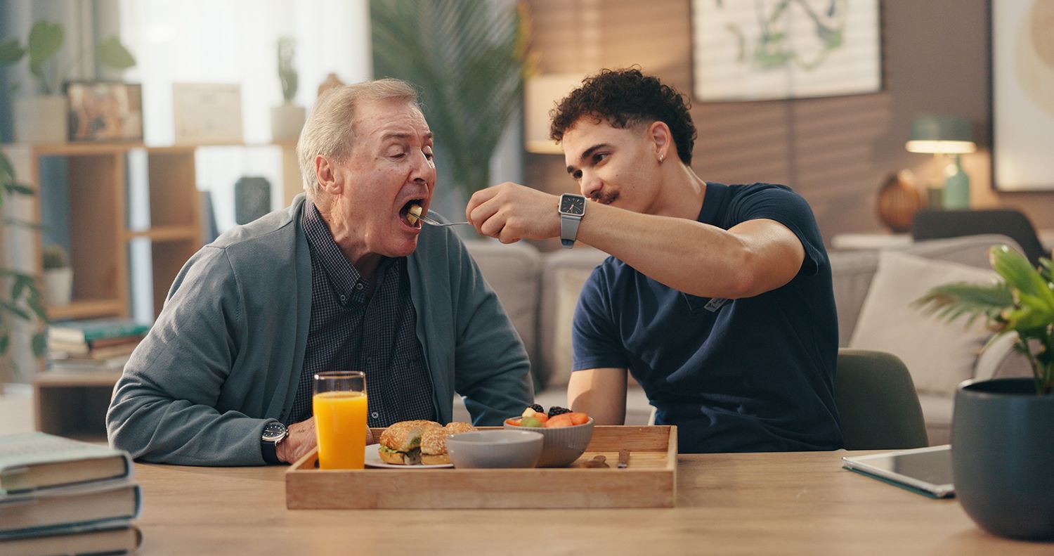 Man feeding an elderly woman at a dining table, with a glass of orange juice and a home kitchen setting.