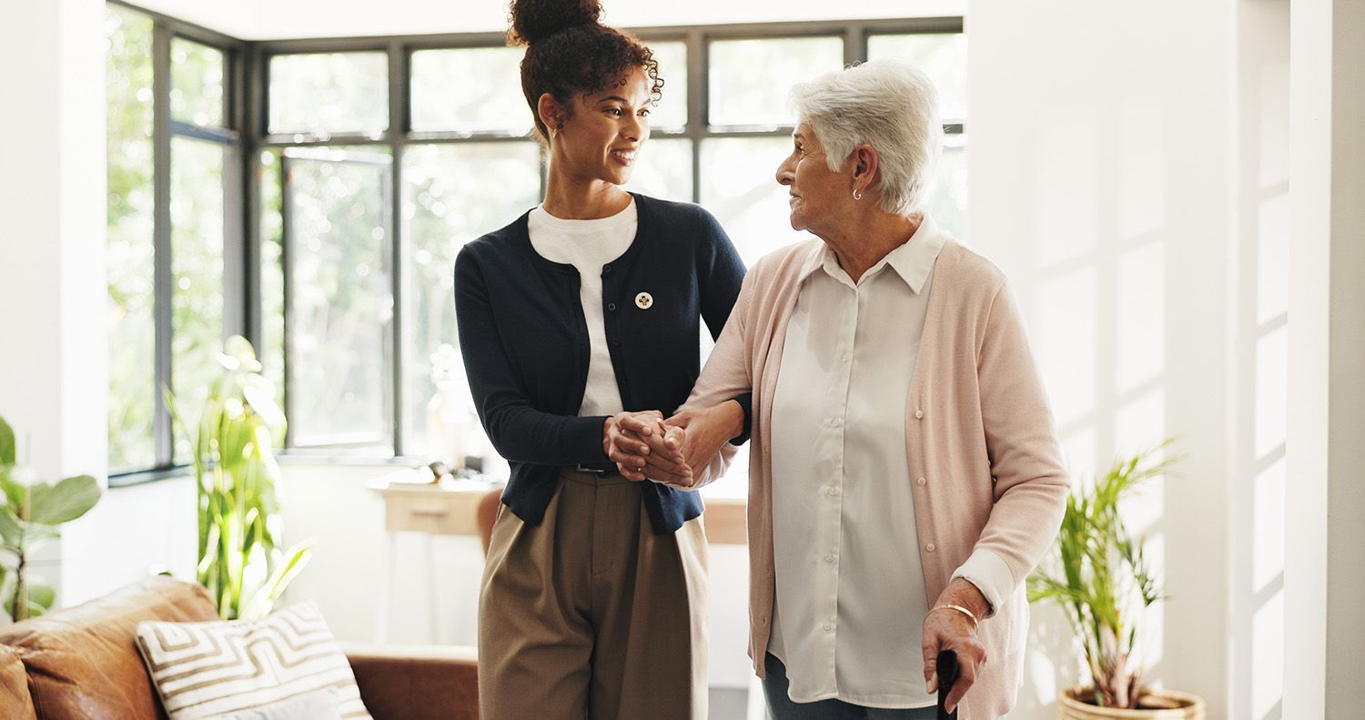 Two women talking in a bright living room, one older, one younger, holding hands and smiling.