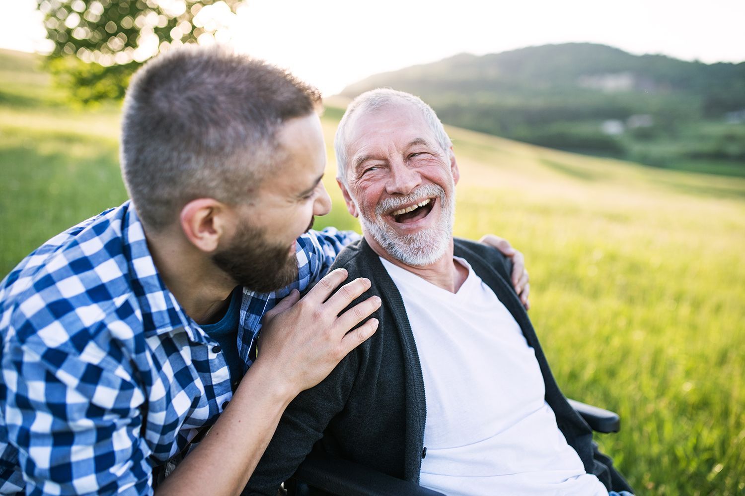 Two people laughing together in a sunny grassy field, one in a blue plaid shirt and the other in a wheelchair