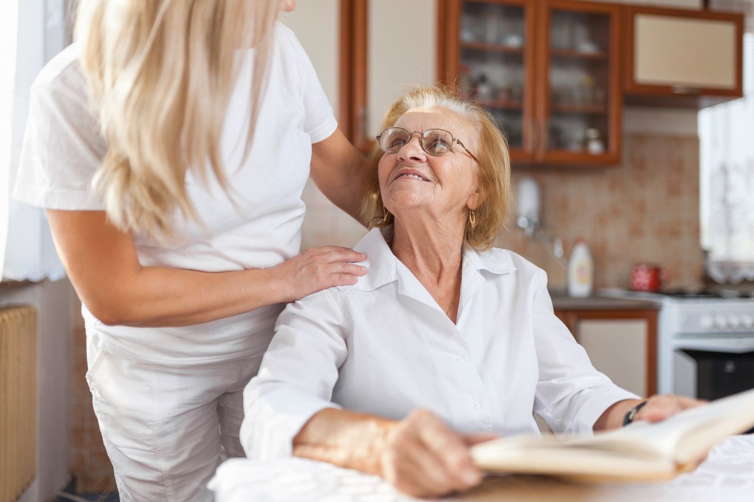 Caregiver standing beside seated woman in a bright kitchen, gently resting a hand on her shoulder