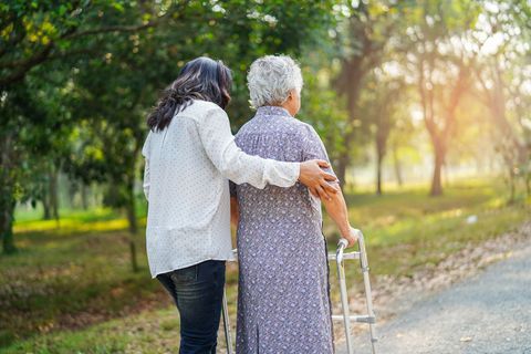 Two people walking outdoors on a sunny path, one helping the other with a walker