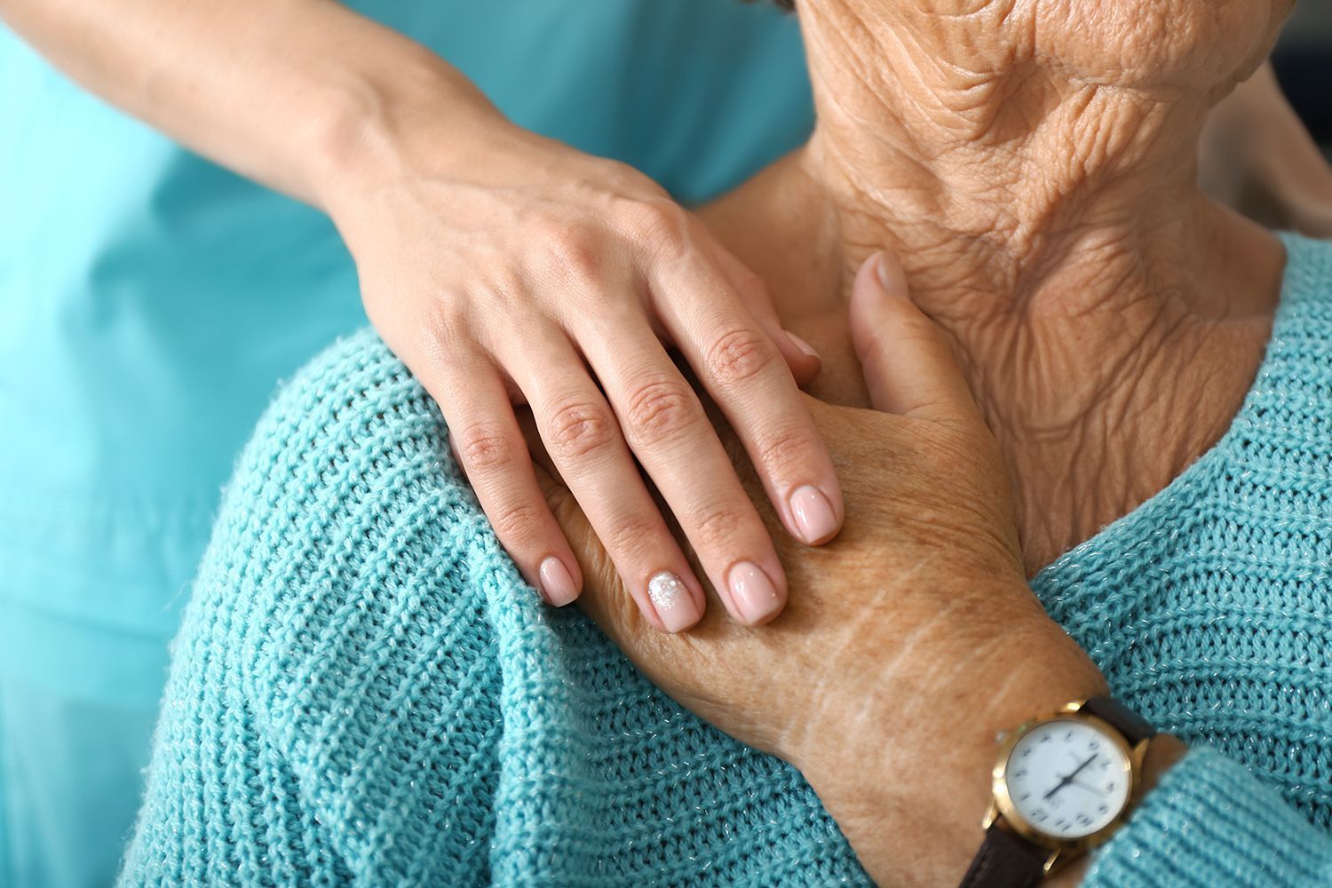 Hands resting on an older person’s shoulder, with a turquoise sweater and watch visible.
