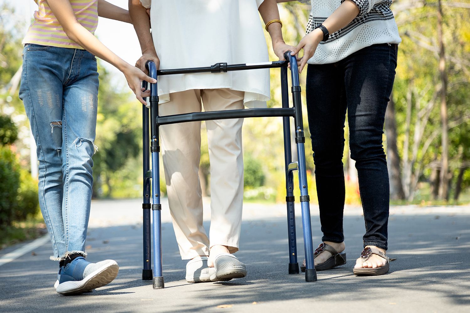 Two people supporting an older person walking with a walker on a sunny road