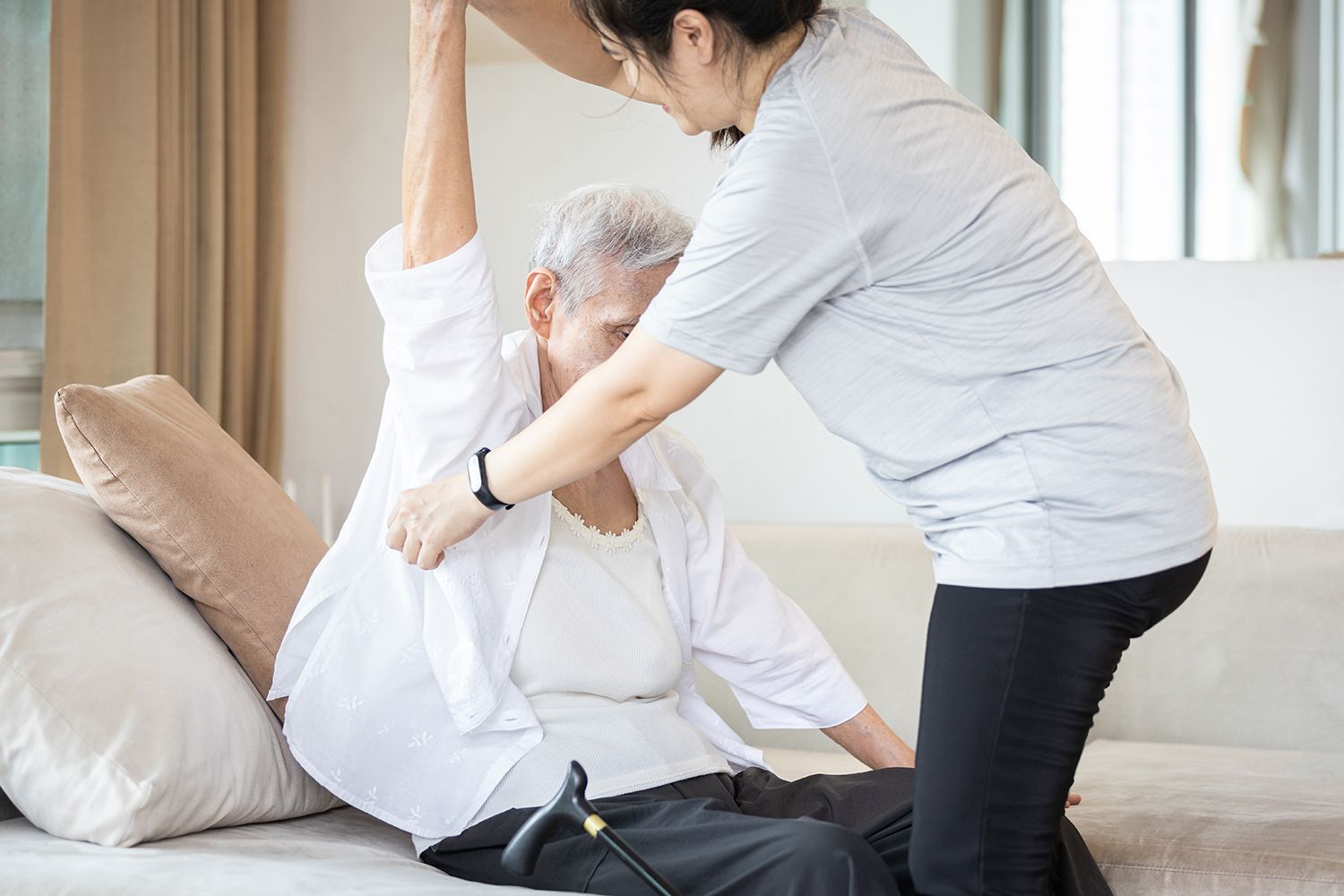 Caregiver helping seated patient stretch arm on a sofa in a bright living room