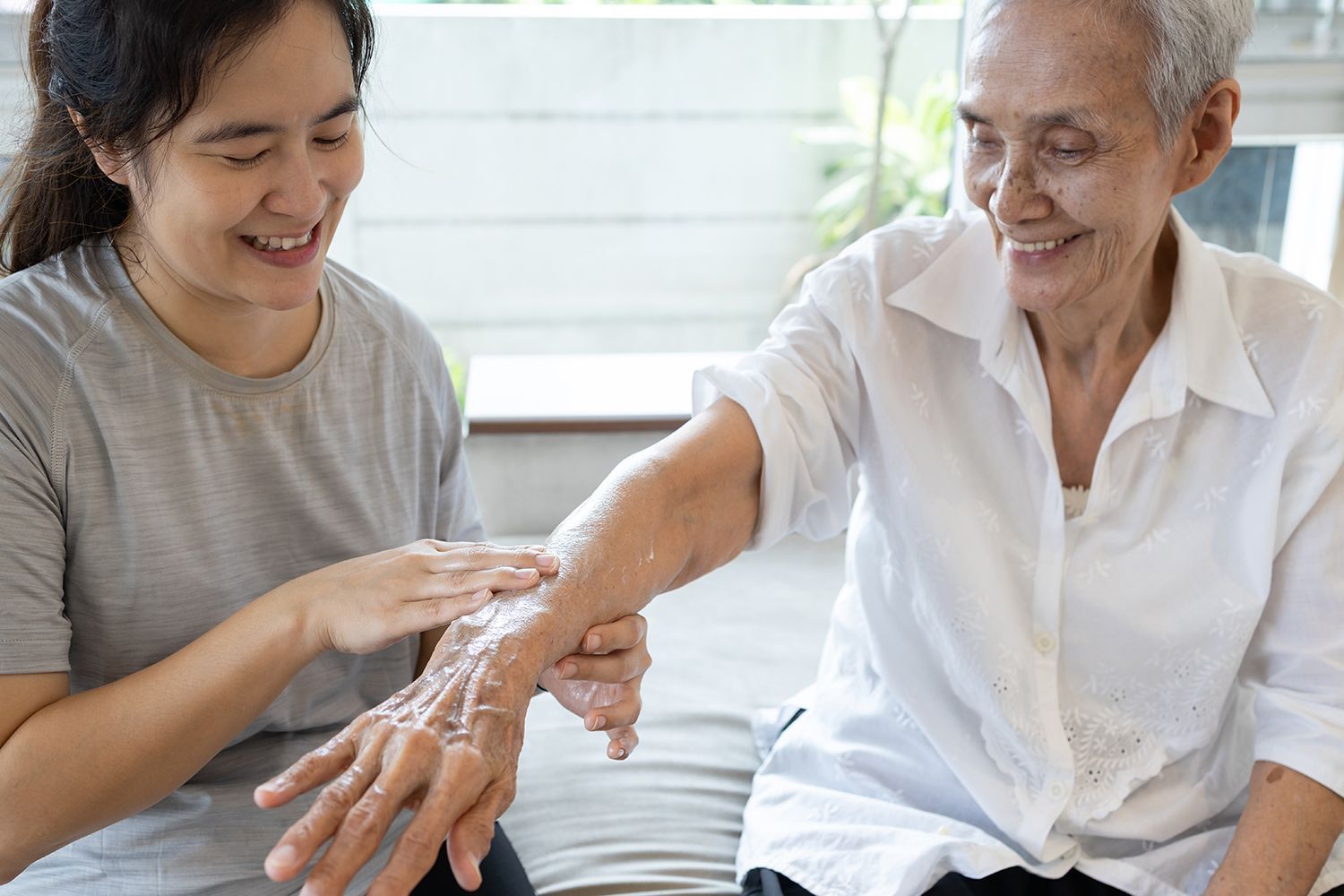 Two women smiling as one gently applies cream to the other’s arm on a couch