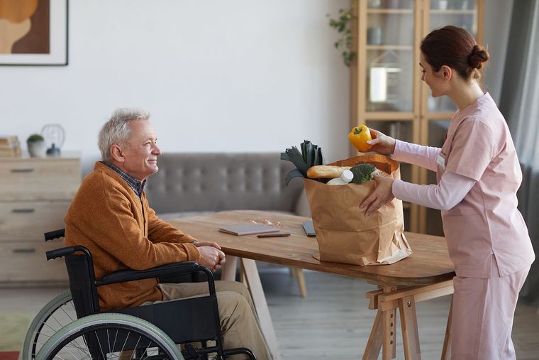 Caretaker hands groceries to man in wheelchair at a dining table