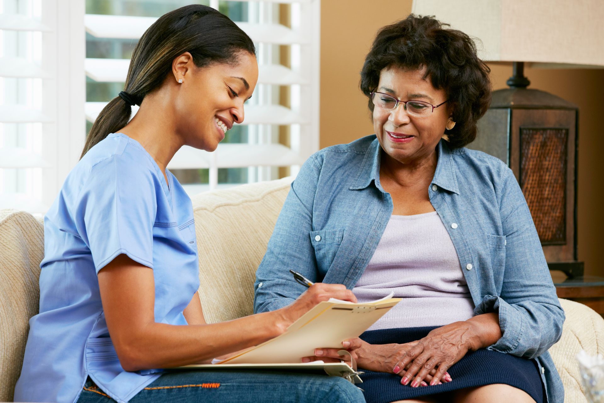 Two women sit on a couch, smiling and reviewing papers together in a bright living room.