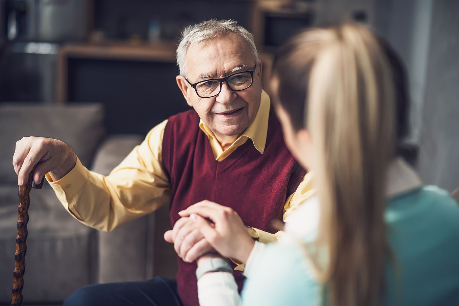 Older man in a burgundy vest speaking with a blonde woman across a table indoors