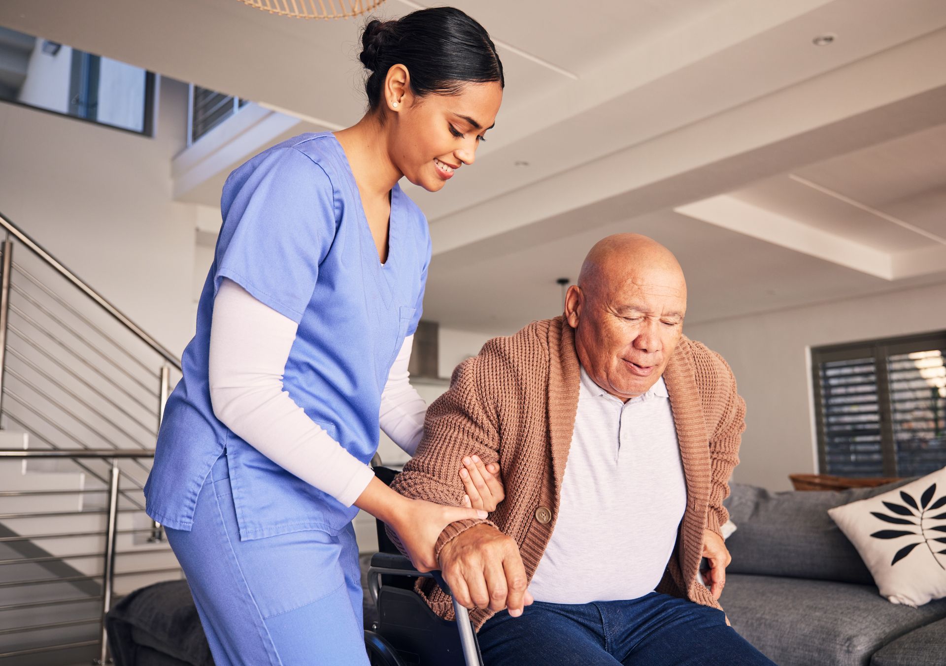 Caregiver in blue scrubs helping an older man stand in a bright living room