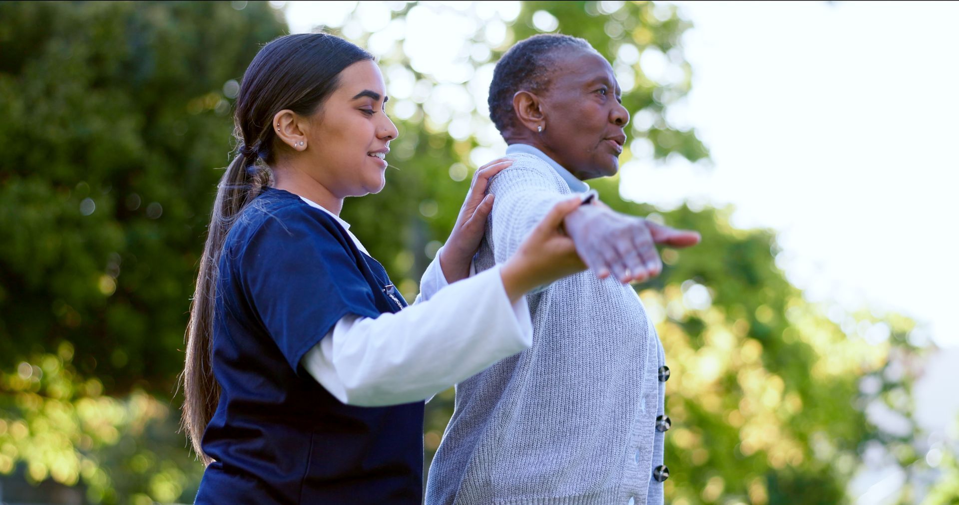 Two people stretching outdoors in a green park, one guiding the other’s arms.