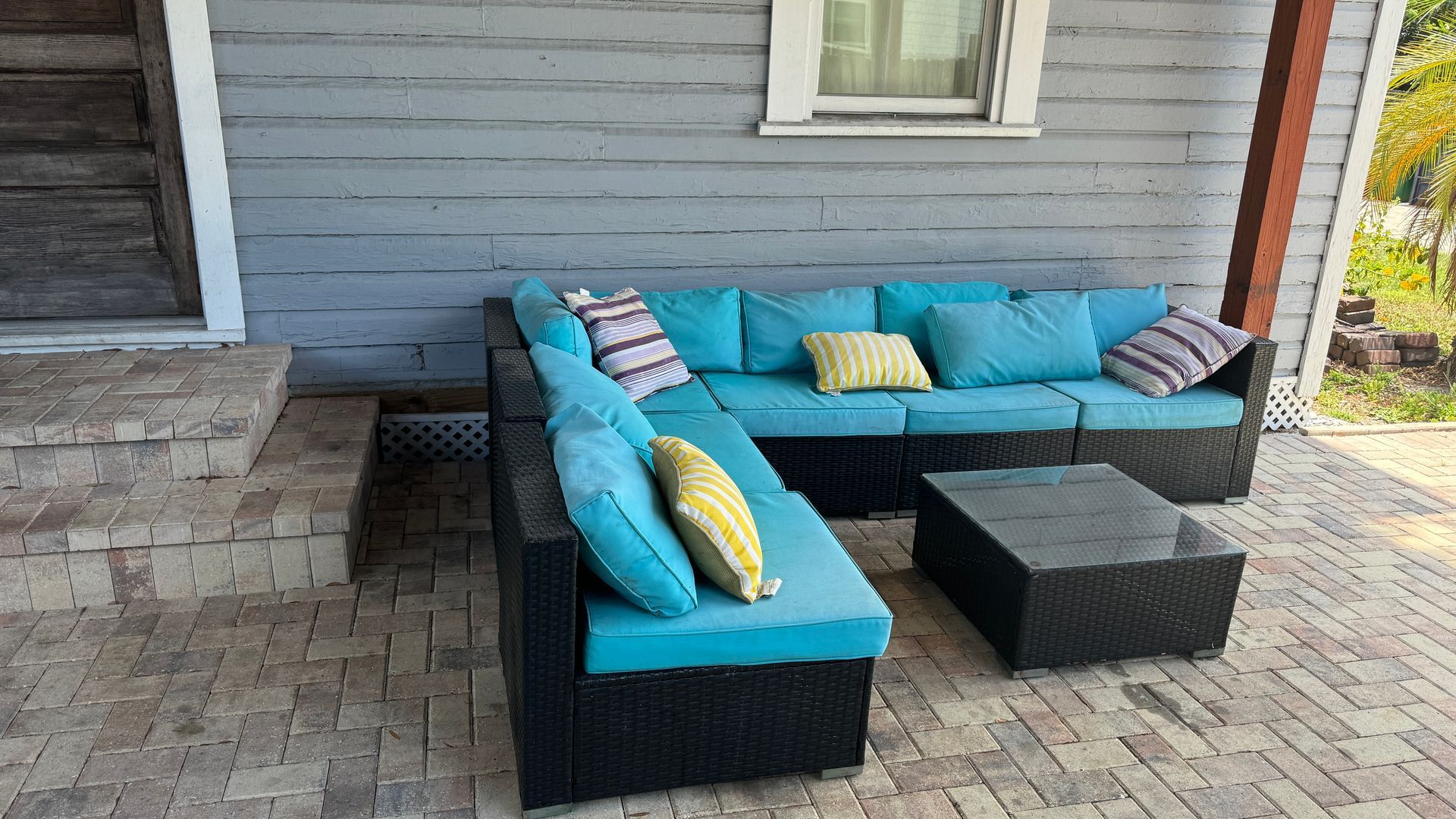 A black couch with blue cushions is sitting on a brick patio in front of a house.