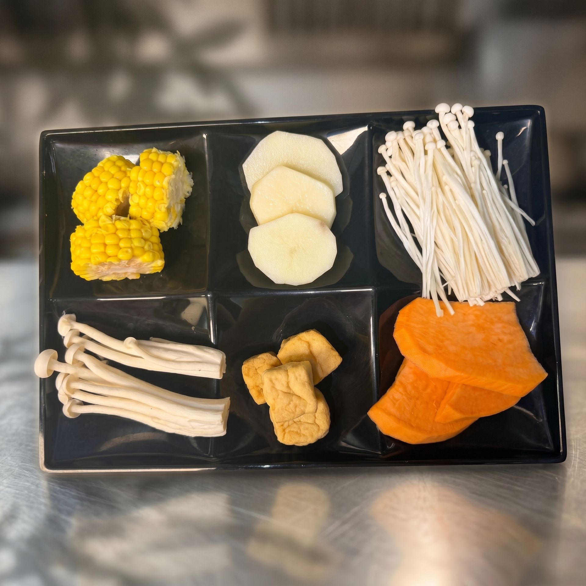 Assorted sliced vegetables arranged in sections on a black tray, including corn, mushrooms, enoki, and pumpkin