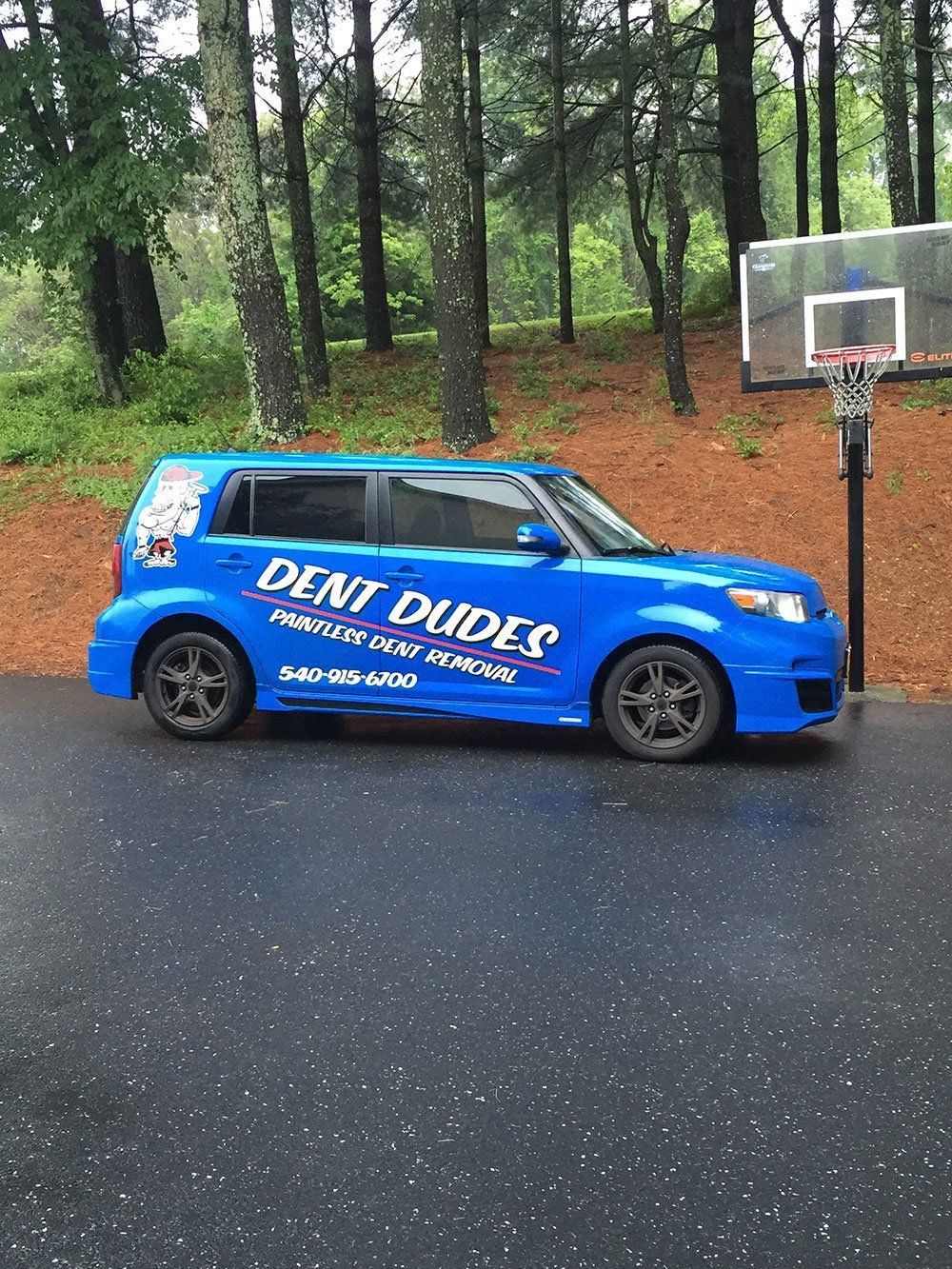 A blue car with dent dudes written on the side is parked next to a basketball hoop.