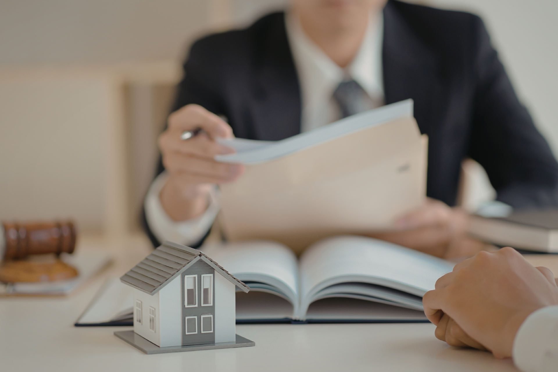 A person in a suit hands papers to another person across a desk, which features a small house model and an open book.