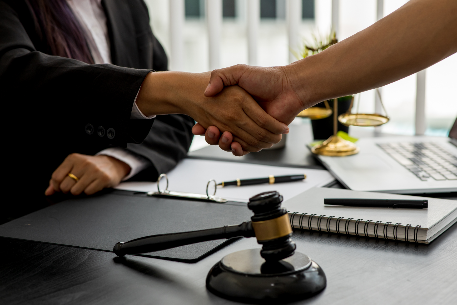 A professional handshake over a desk with a wooden gavel, notebook, and scales of justice in the background.