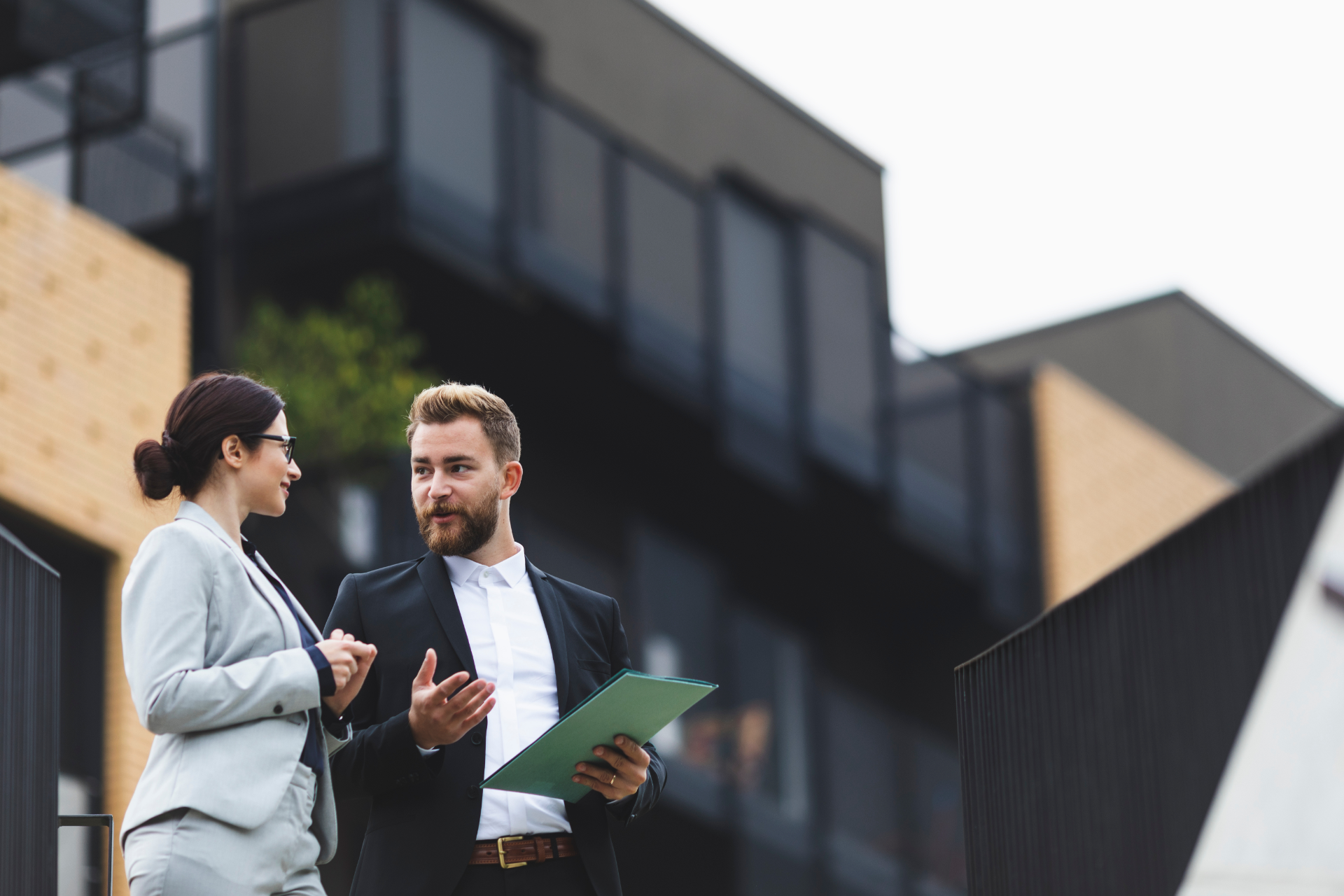 Two professionals in business attire stand outside a modern building, talking while one holds a green folder.