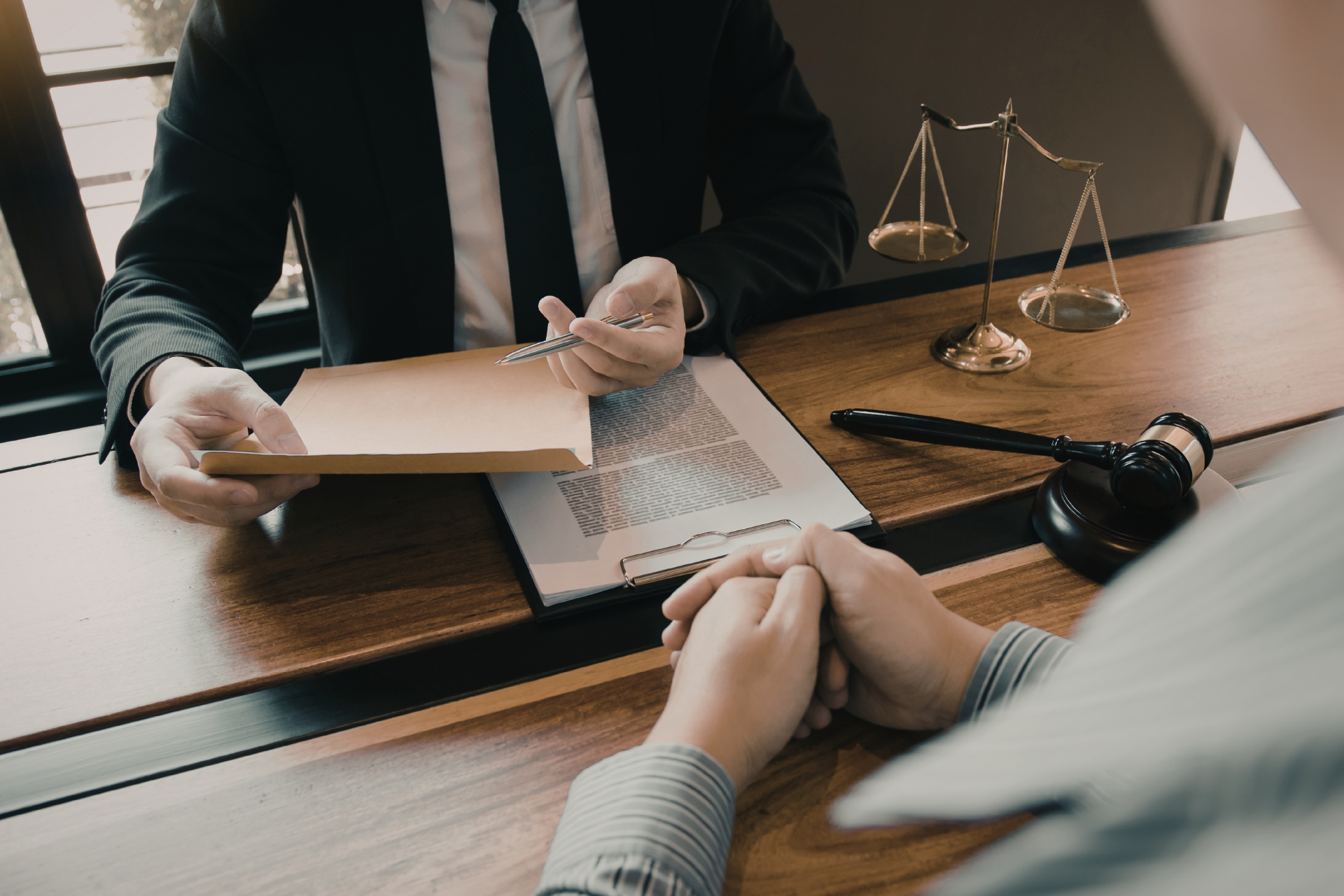 A person in a suit hands a document to another person across a desk with a scale of justice and a judge's gavel.