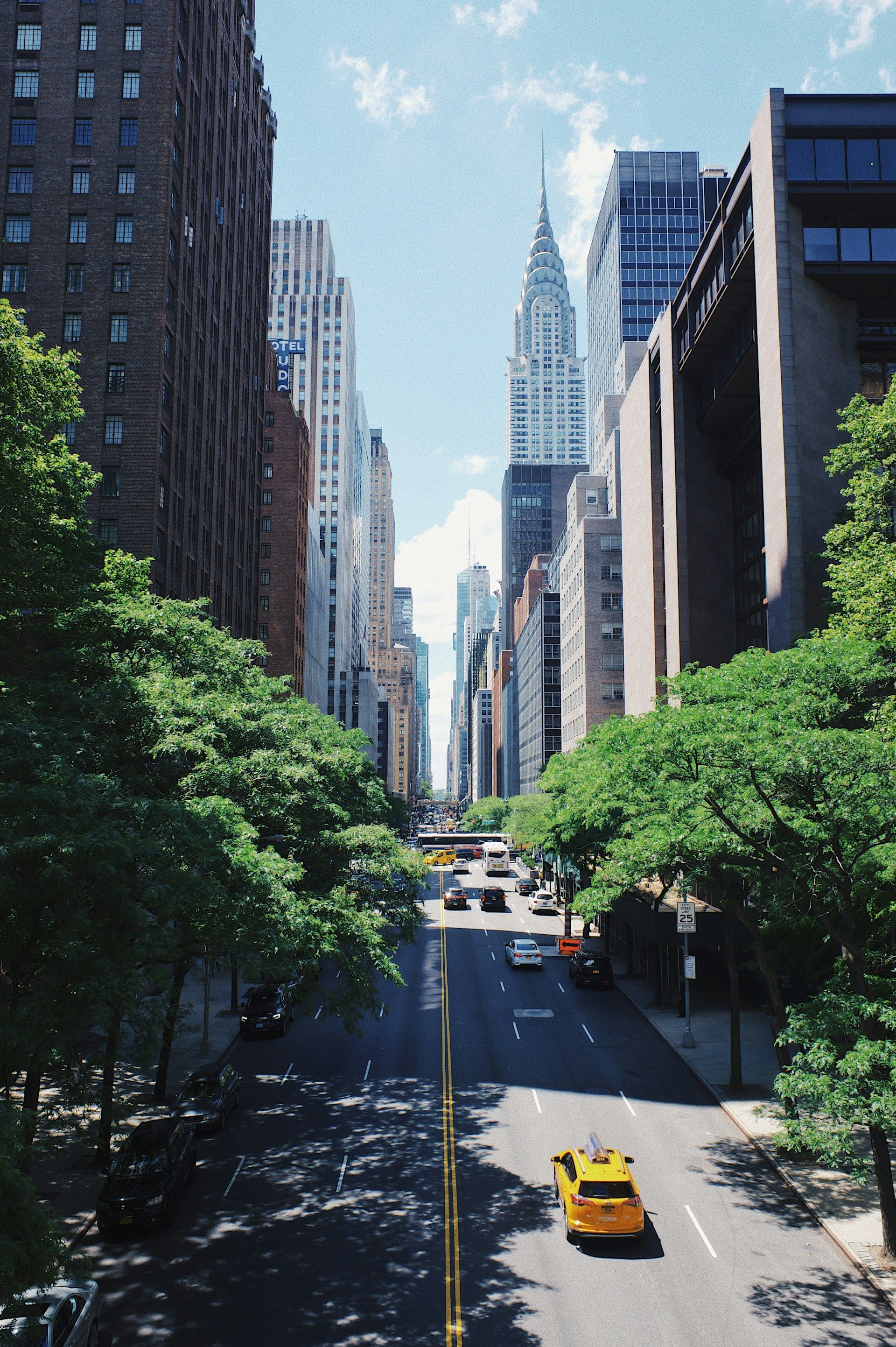 A sunny New York City street lined with green trees and skyscrapers, featuring a yellow taxi driving on the road.