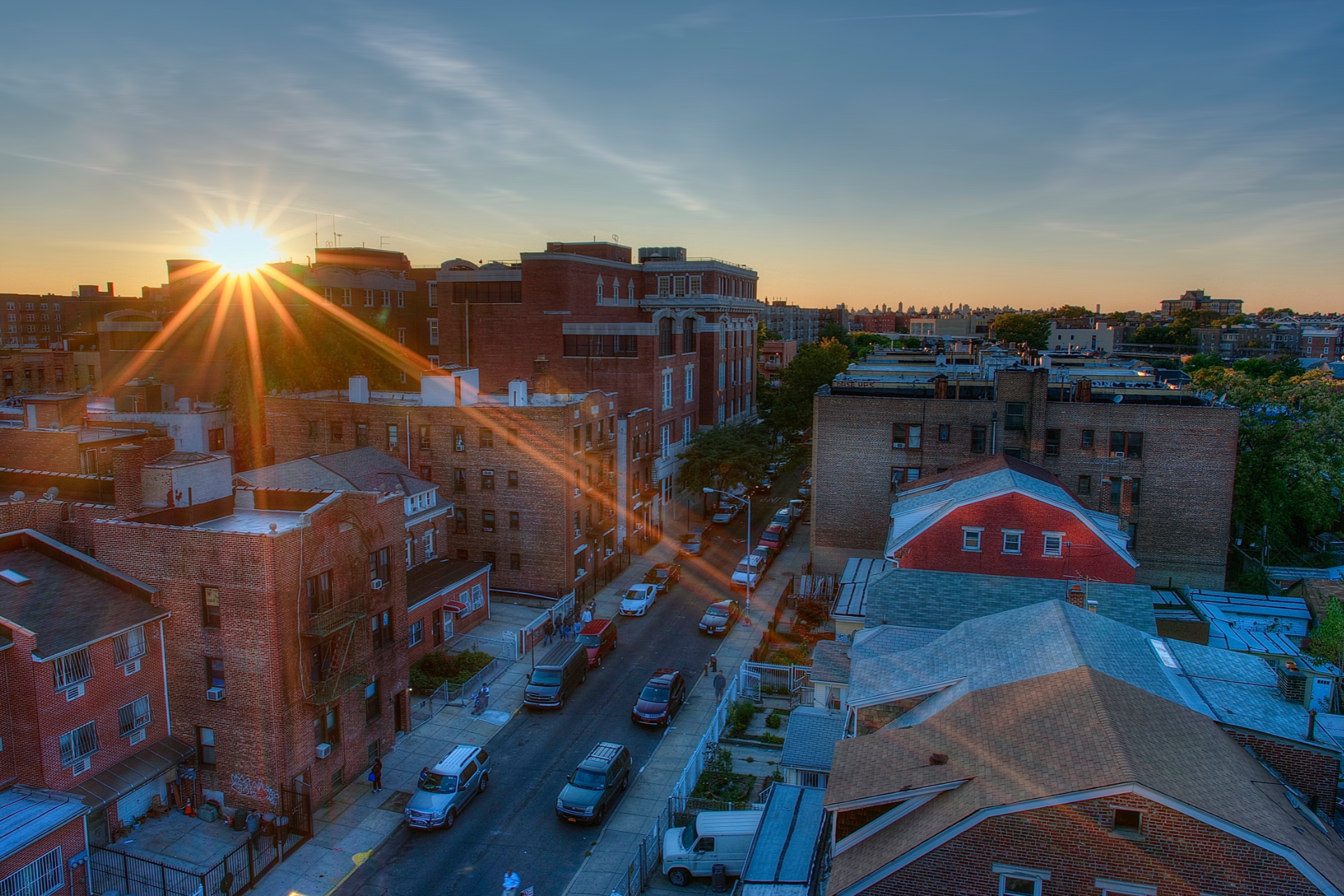 Sunset over a city street with brick buildings, parked cars, and a red-roofed structure.