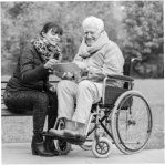 Woman handing a mug to an elderly person in a wheelchair outdoors