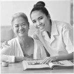 Two women smiling at a table with an open book, one seated and one leaning close in a bright room