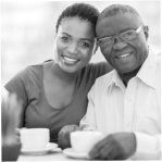 Smiling couple seated at a table with coffee cups, posing closely together in a bright setting