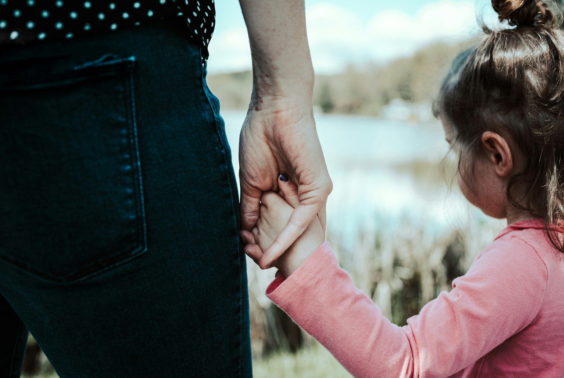 Adult and child holding hands outdoors near water, with the child in a pink sweater