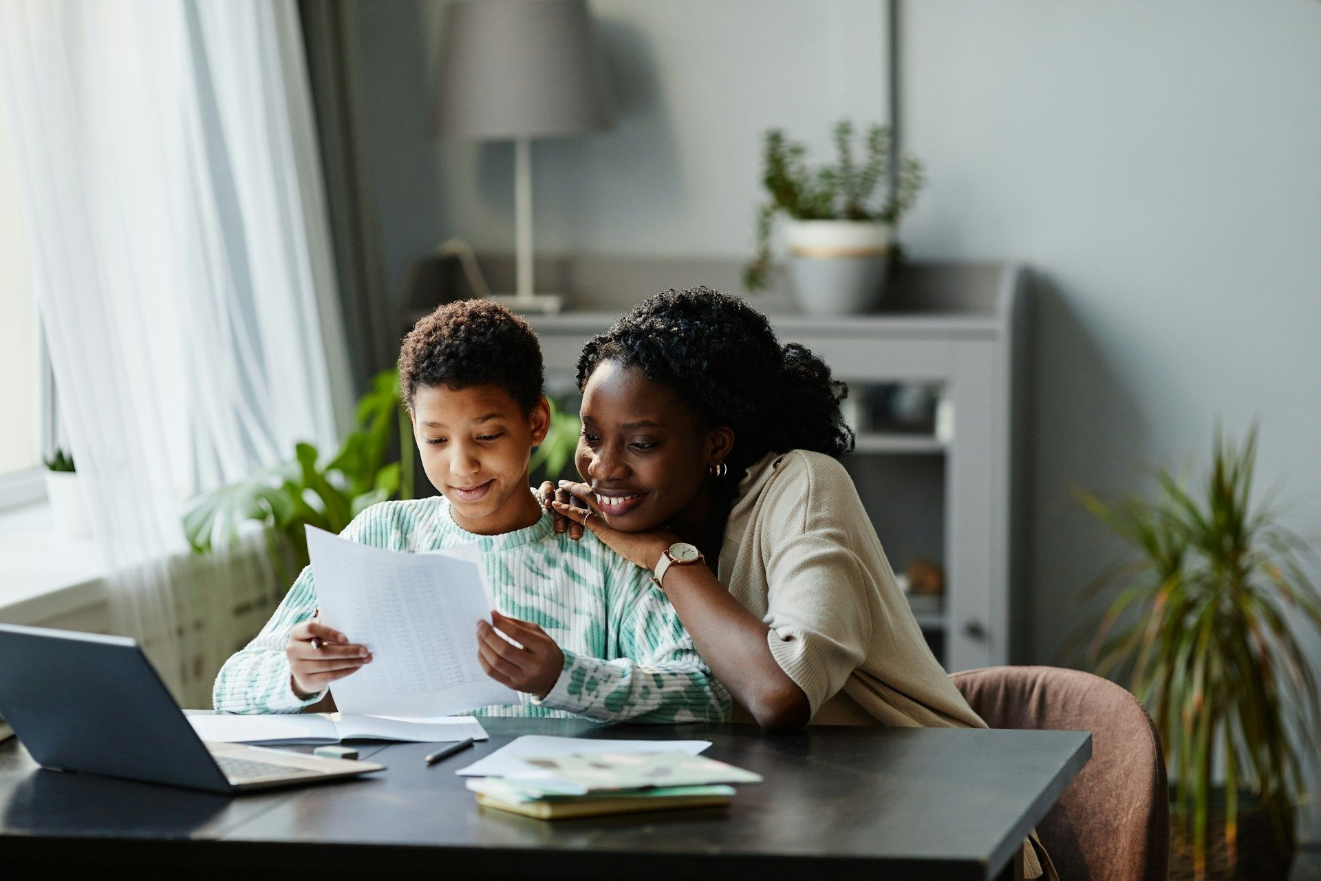 Adult helping child read papers at a table with a laptop in a bright home office