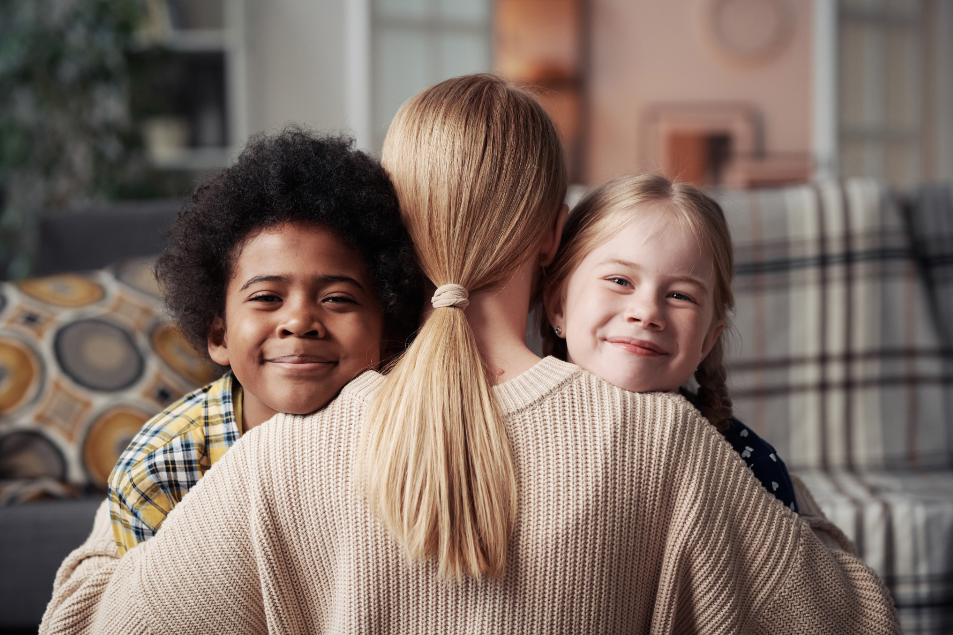 Two smiling children hug a person seated on a couch in a cozy living room.