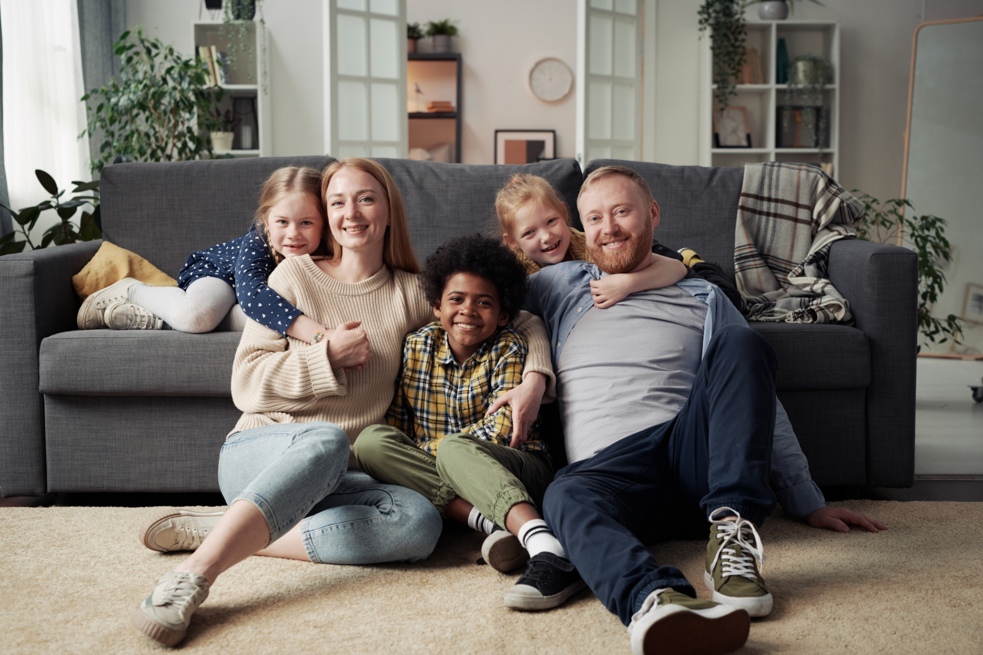 Four people smiling on a living room floor in front of a gray sofa, with two dogs nearby
