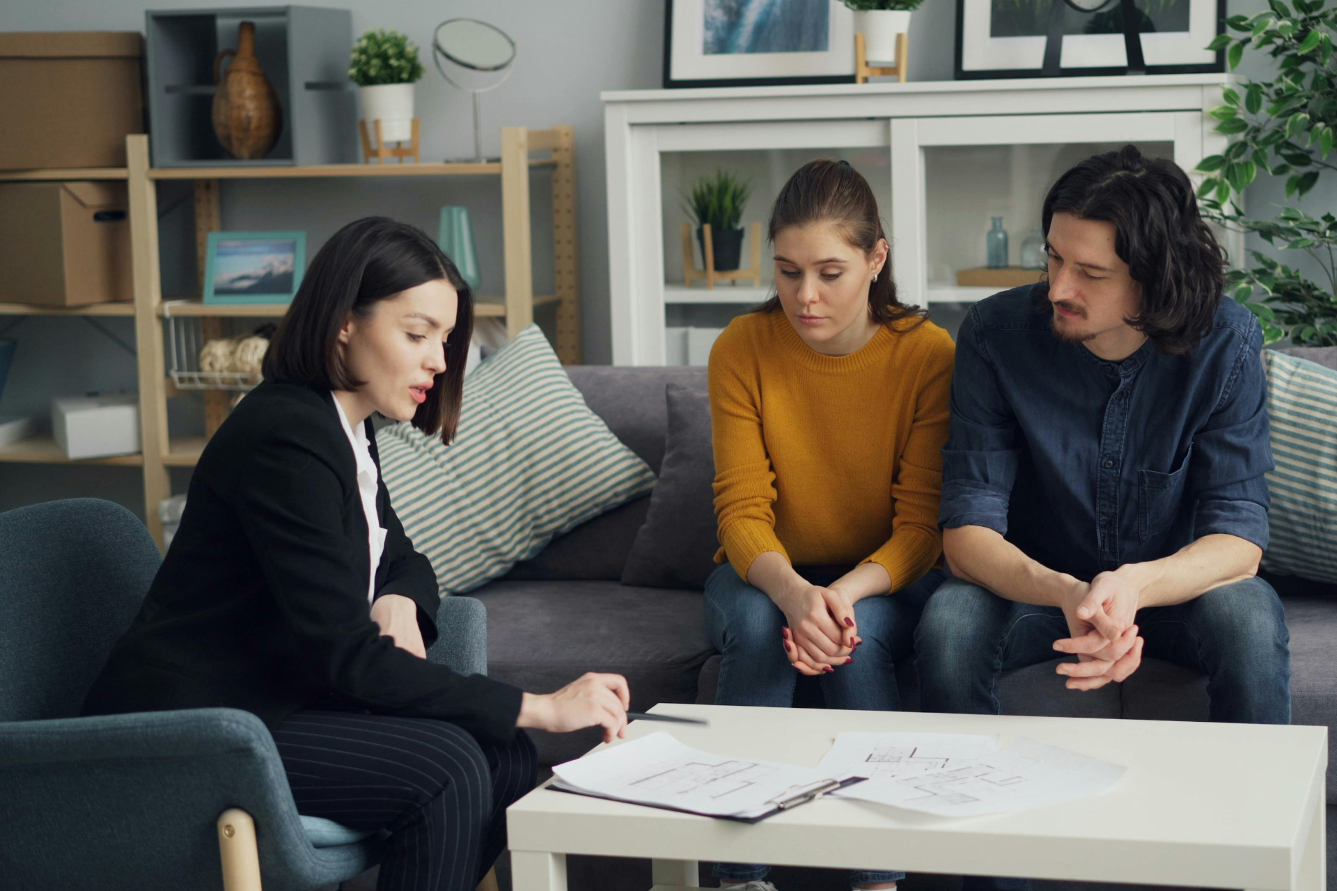 Three people reviewing papers around a white coffee table in a living room, with shelves and decor in the background