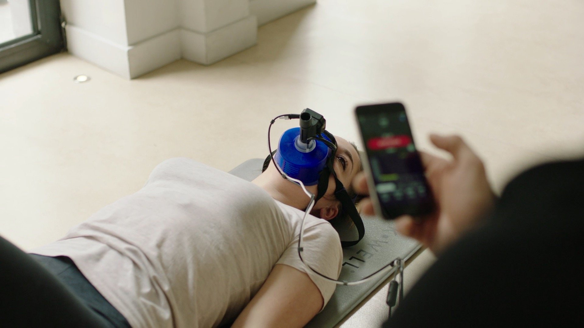 Person with blue respiratory mask on bed, observed by someone holding smartphone.