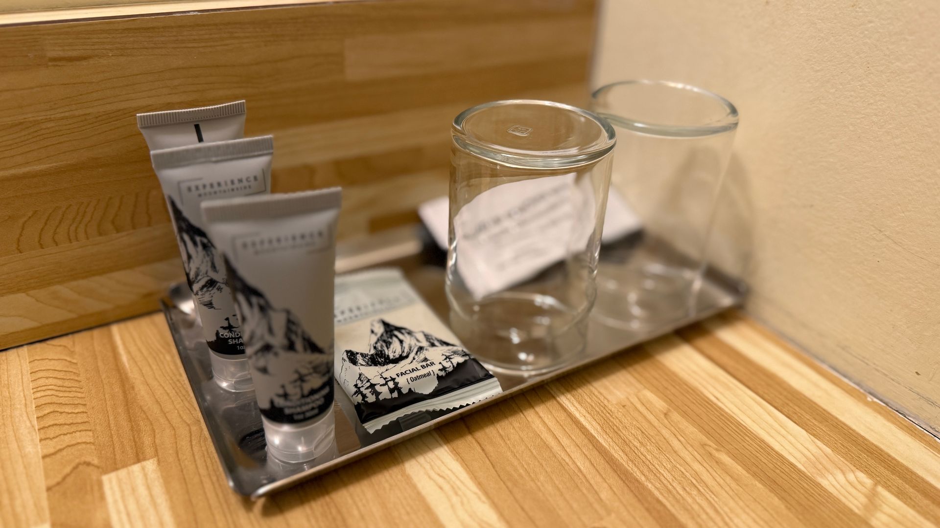 A metal tray on wooden surface holding toiletries: two clear glasses, three tubes, a card, near a wall.