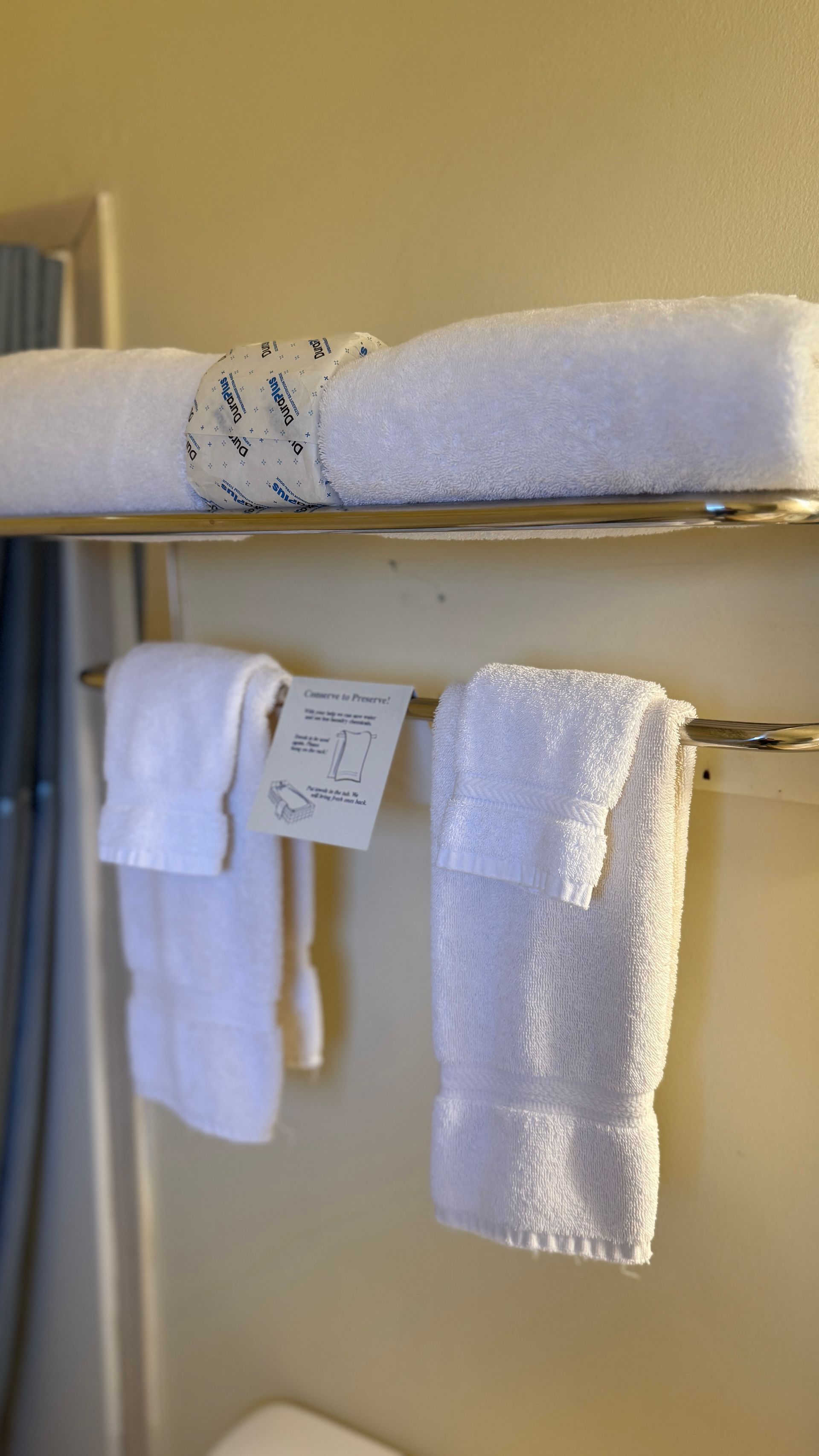 White towels on a chrome rack in a bathroom, one rolled and banded, two folded with a handwritten note.